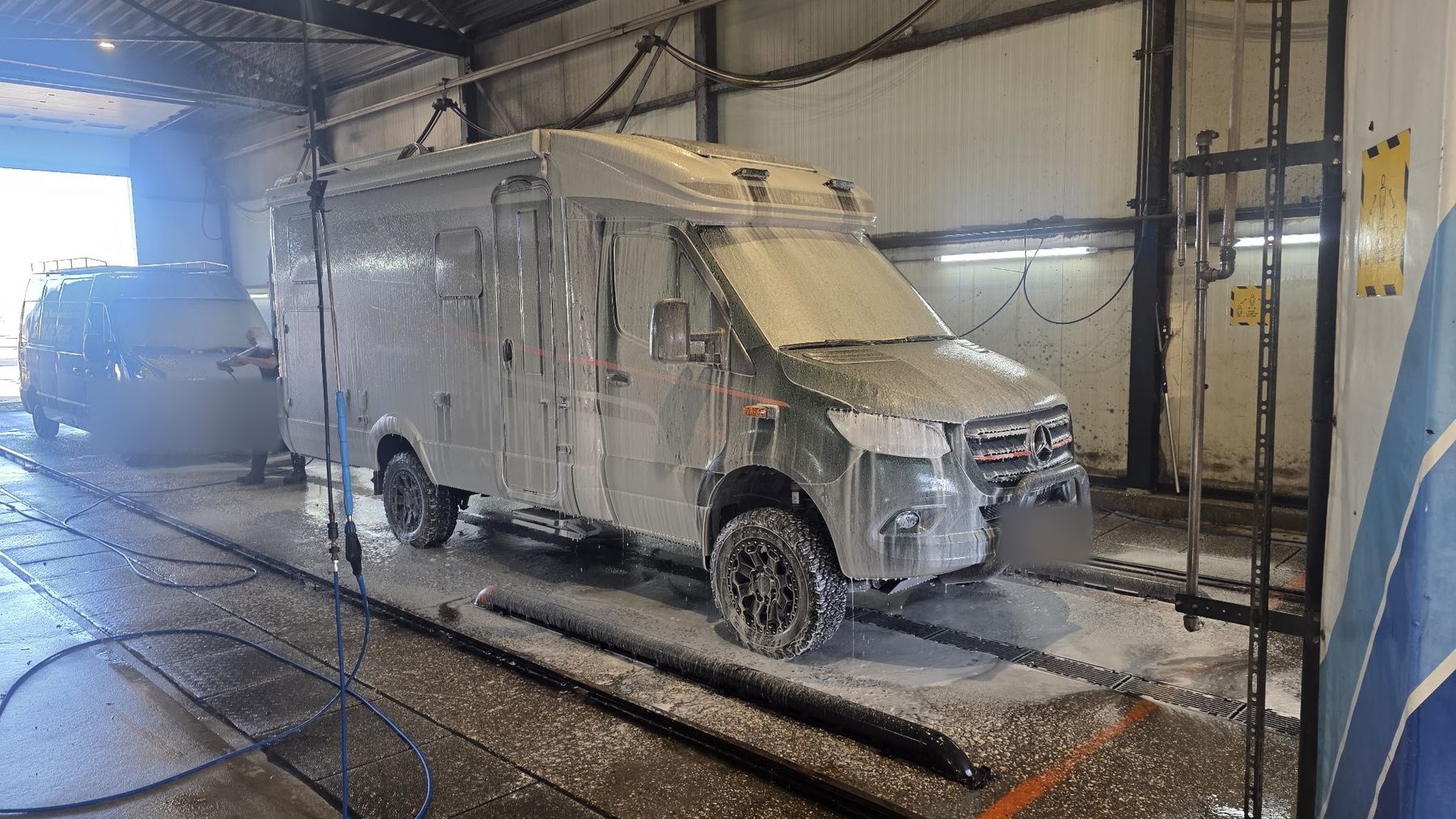 Een camper onder het schuim in de wasstraat, met een medewerker met een hogedrukspuit op de achtergrond